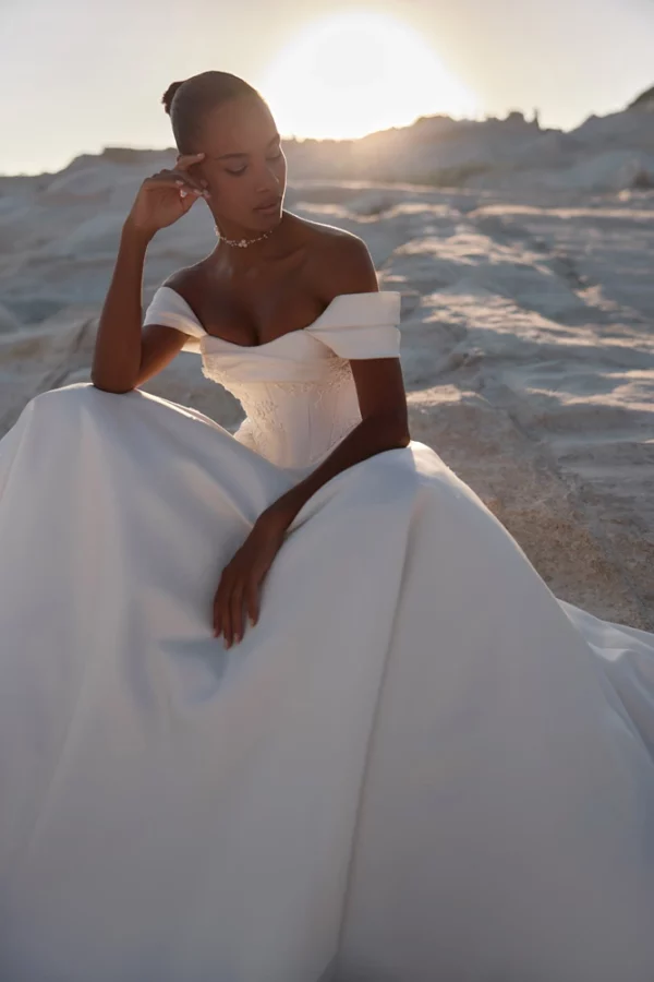 Bride seated in Eva Lendel Chabli wedding gown, showcasing soft satin skirt and off-shoulder neckline at sunset